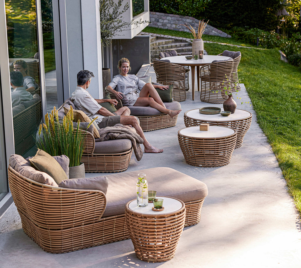 Twee personen ontspannen op geweven meubels omringd door tafels en groen, terwijl ze genieten van een casual buitensetting.