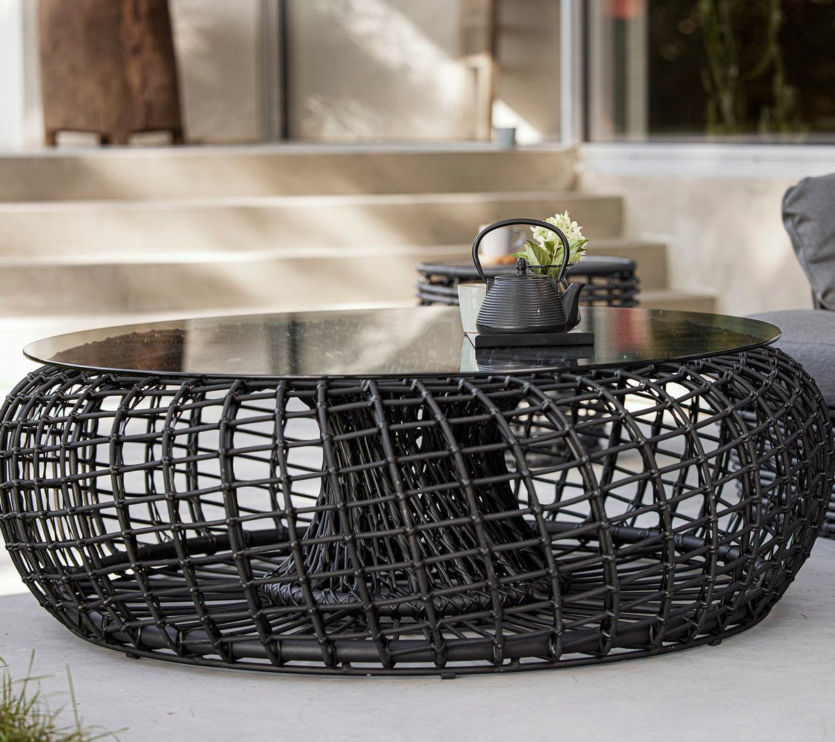 Een modern, rond salontafel met een geweven zwarte basis en een glazen blad, met decoratieve items zoals een plant en een kleine kom.