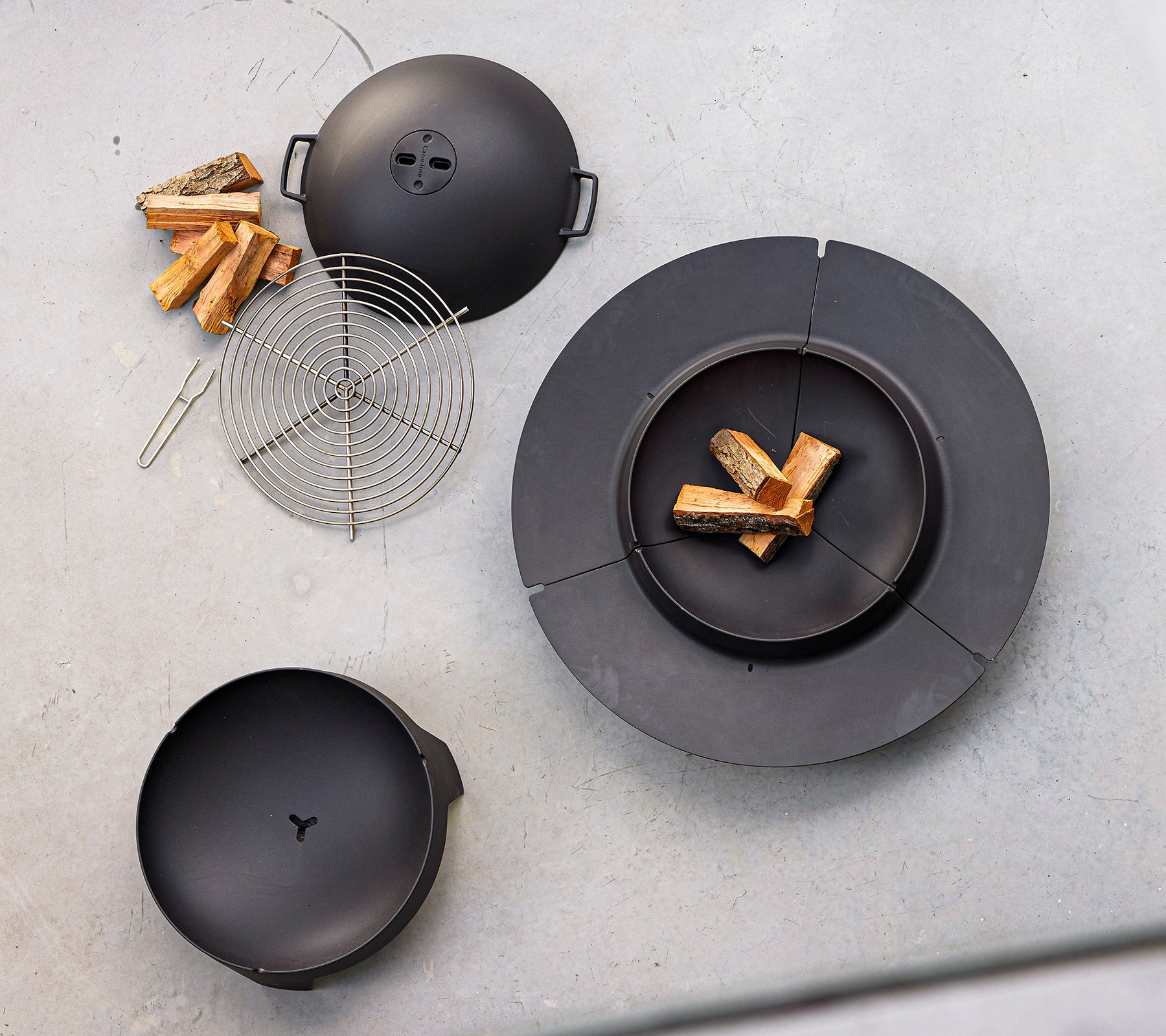 Zwart kookgerei neergelegd met brandhout en een draadgrillrek, ontworpen voor buiten grillen en koken boven vuur.