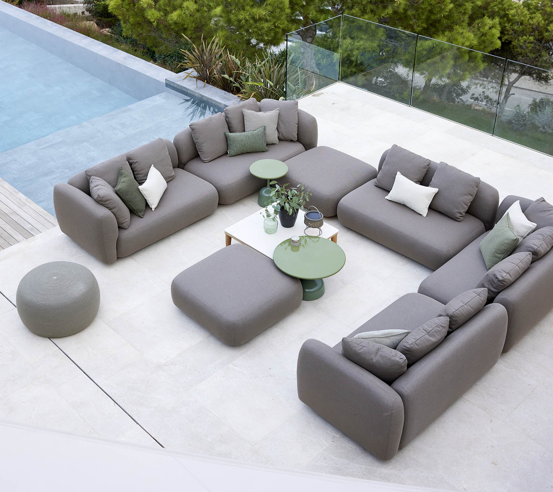 Moderne buitenzitplaatsen met grijze sofa's en groene tafels naast een zwembad, omgeven door groen.