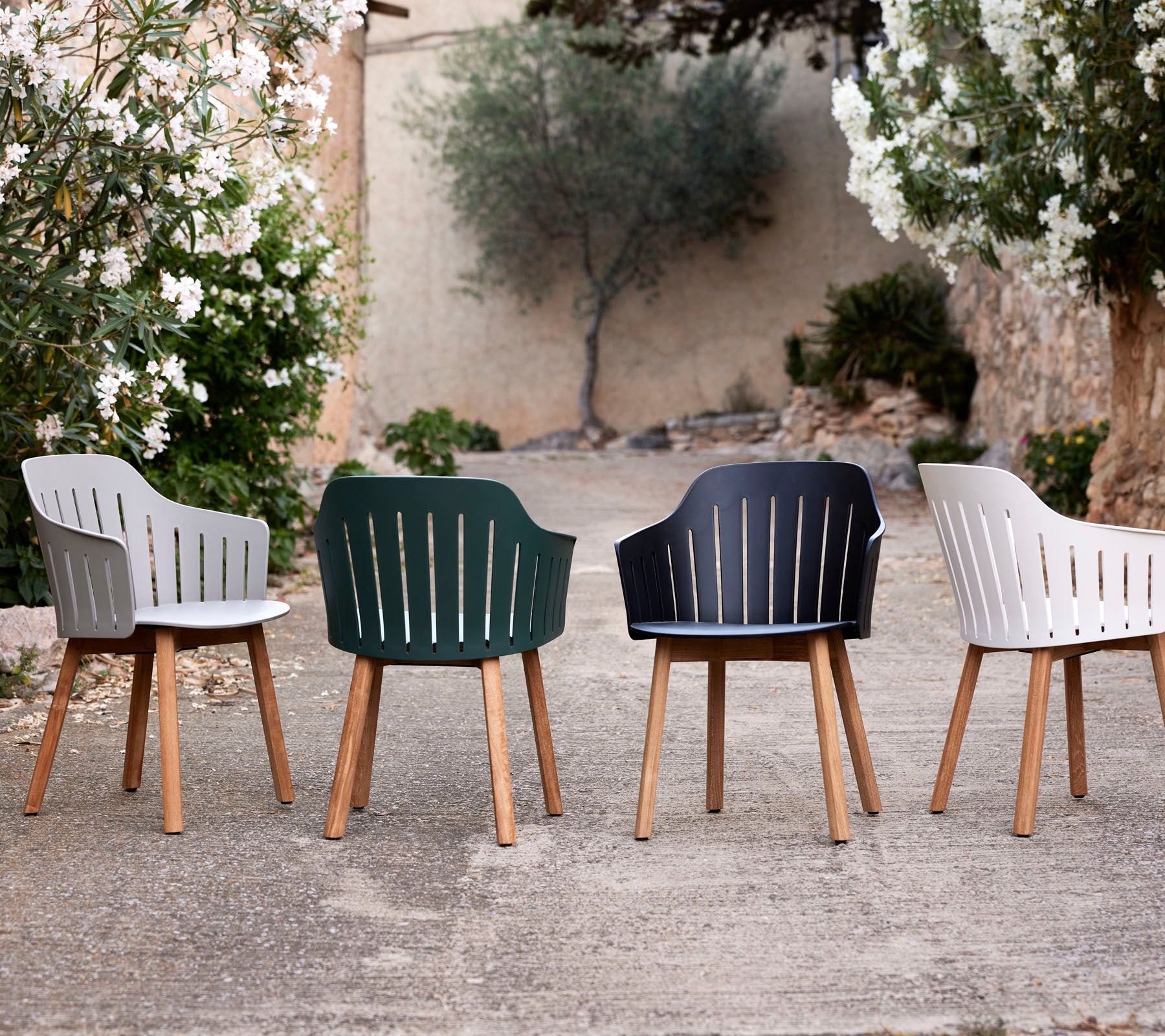 Vier stijlvolle stoelen in verschillende kleuren met houten poten, die een modern ontwerp en lattenrugleuning laten zien temidden van groen.