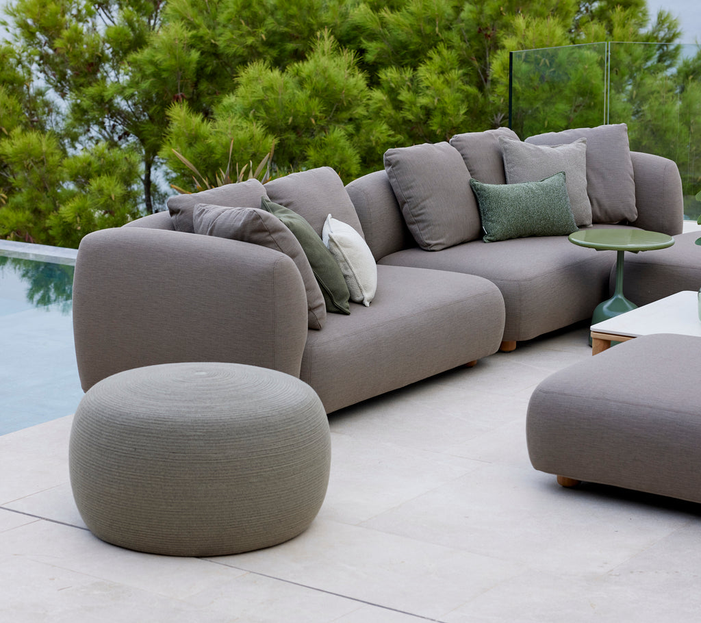 Grote taupekleurige cirkel voetenbank naast een moderne sofa met groene kussens, omgeven door weelderig groen en een serene sfeer.