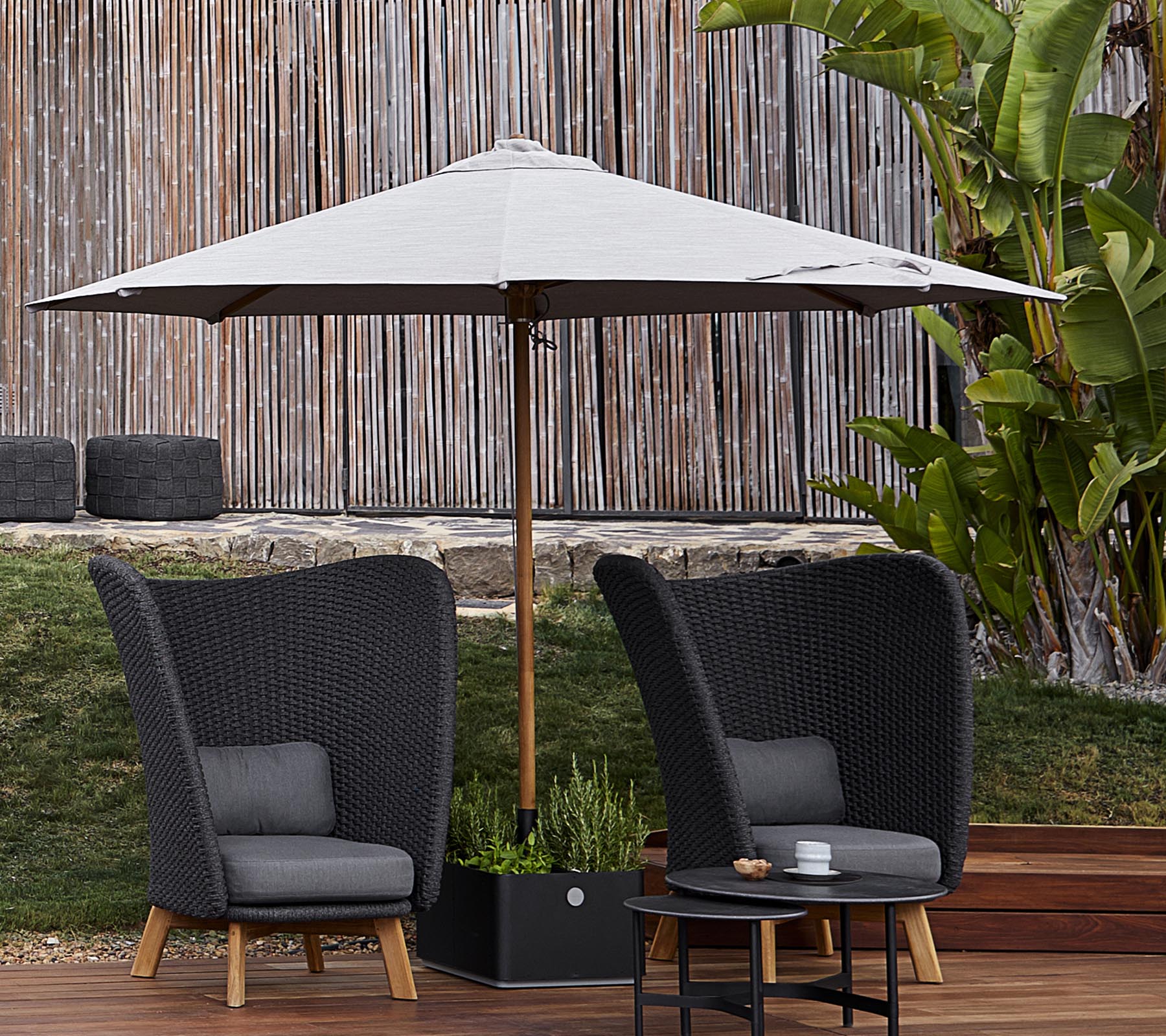 Twee stijlvolle gevlochten stoelen met kussens staan onder een grote parasol, vergezeld van een kleine tafel, tegen een weelderige groene achtergrond.