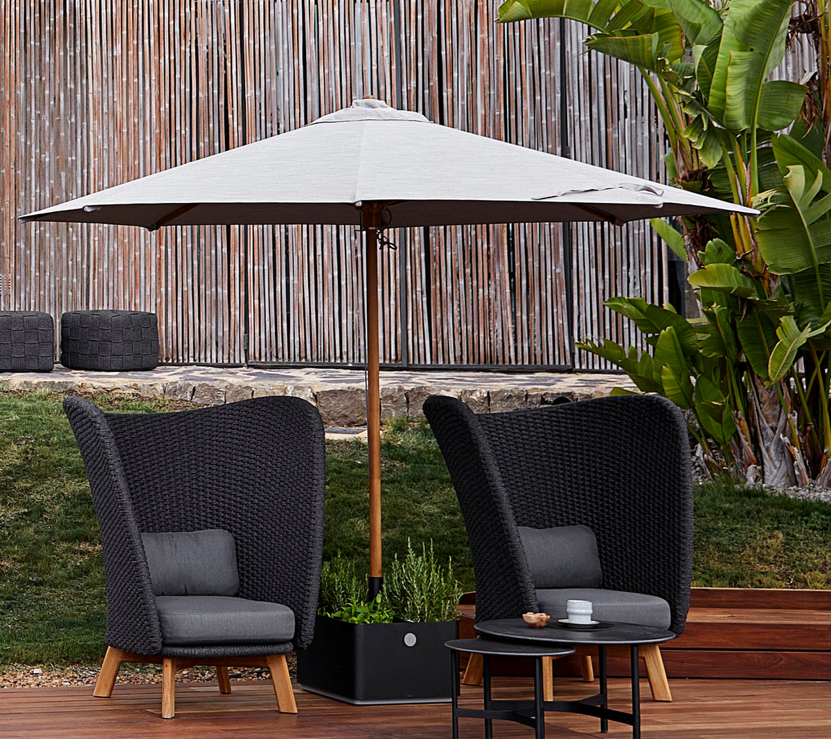 Twee stijlvolle gevlochten stoelen met kussens staan onder een grote parasol, vergezeld van een kleine tafel, tegen een weelderige groene achtergrond.
