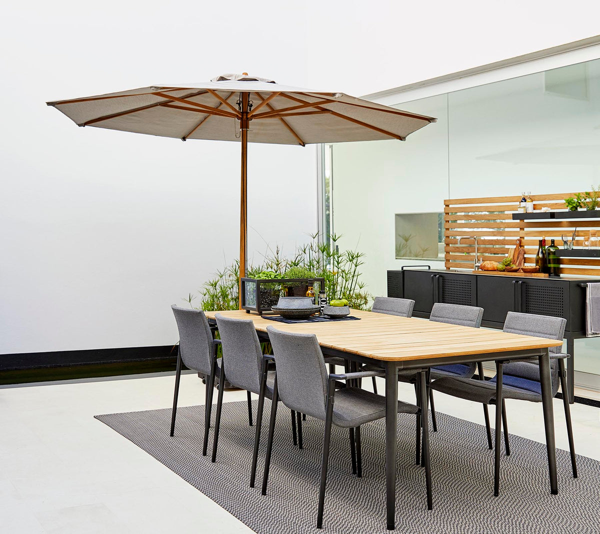 Een klassiek eetset met een houten tafel met een parasol en grijze stoelen, omringd door decoratieve planten op een stijlvolle vloerbedekking.