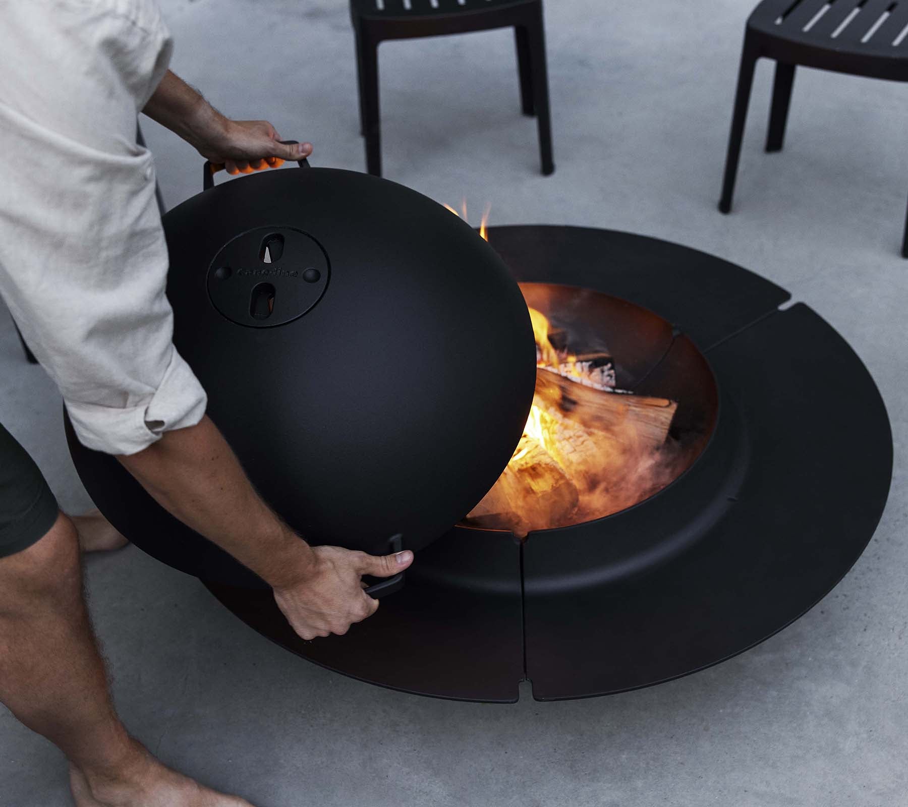 Een persoon past een deksel aan op een slanke zwarte Ember vuurkorf met vlammen en brandhout zichtbaar binnenin, omringd door stoelen.