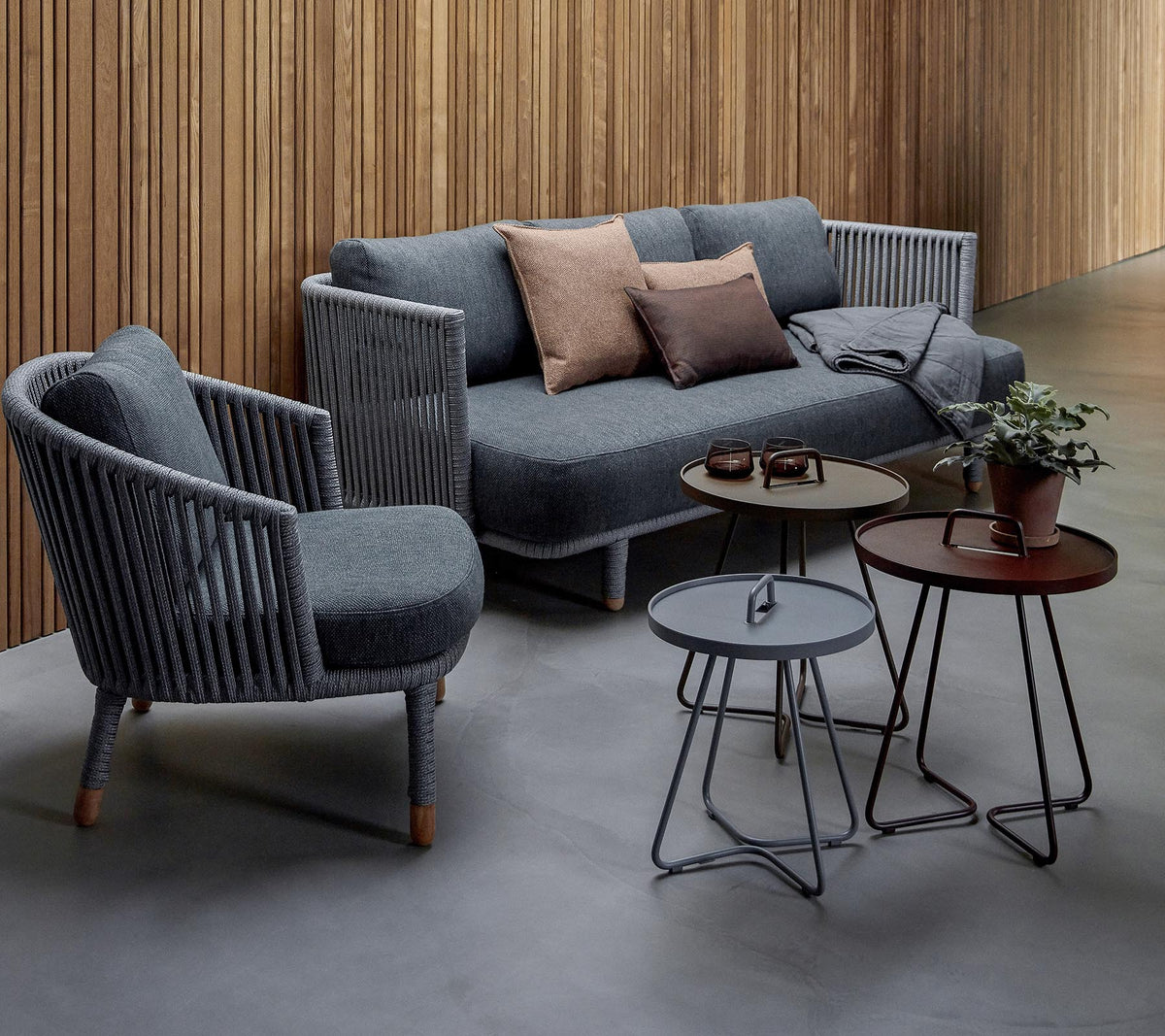 Stijlvolle zithoek met een grijze bank met kussens en twee accentstoelen, vergezeld van drie moderne tafels en een potplant.