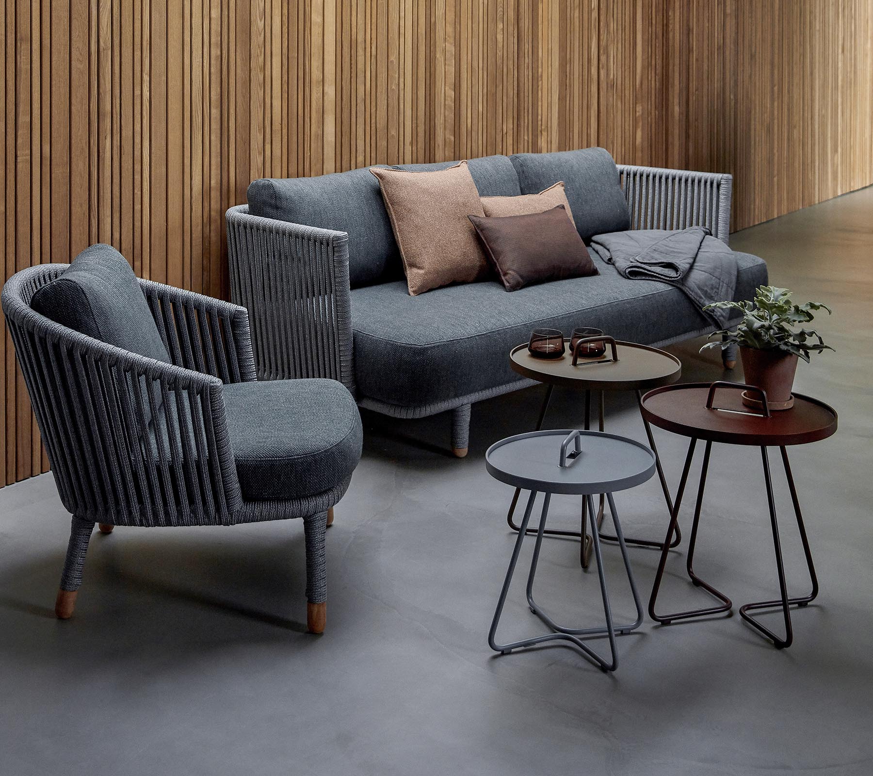 Stijlvolle zithoek met een grijze bank met kussens en twee accentstoelen, vergezeld van drie moderne tafels en een potplant.