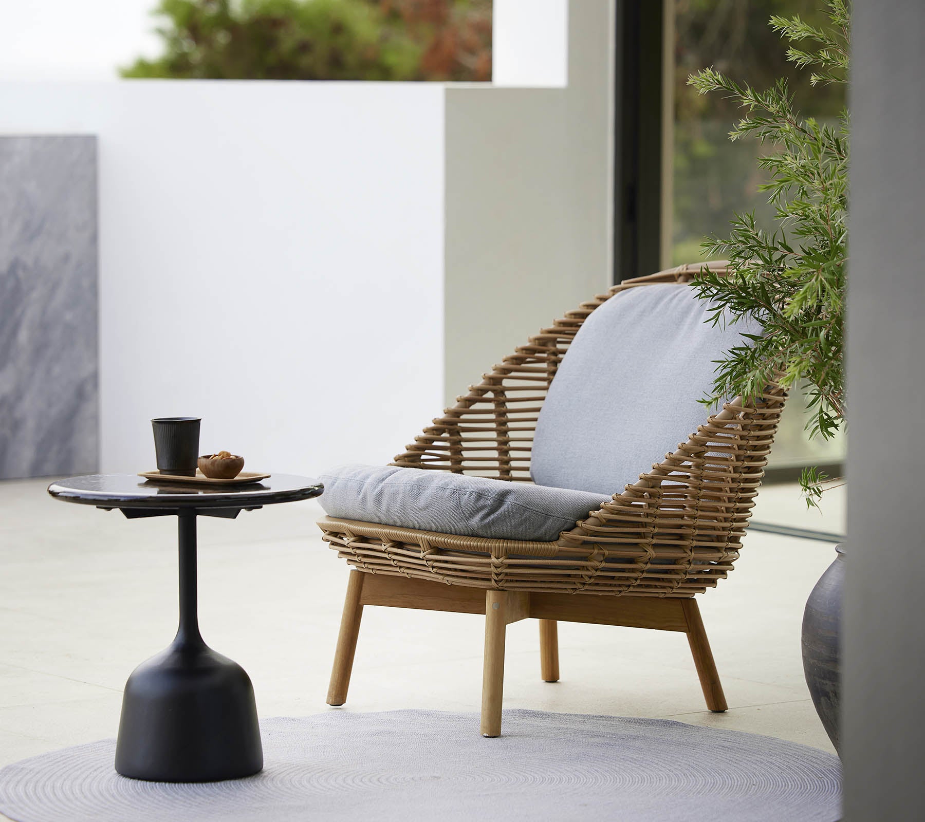 Een stijlvolle loungestoel met een geweven ontwerp en zachte kussens, vergezeld van een slank bijzettafeltje en een kleine plant. Ideaal voor ontspanning.