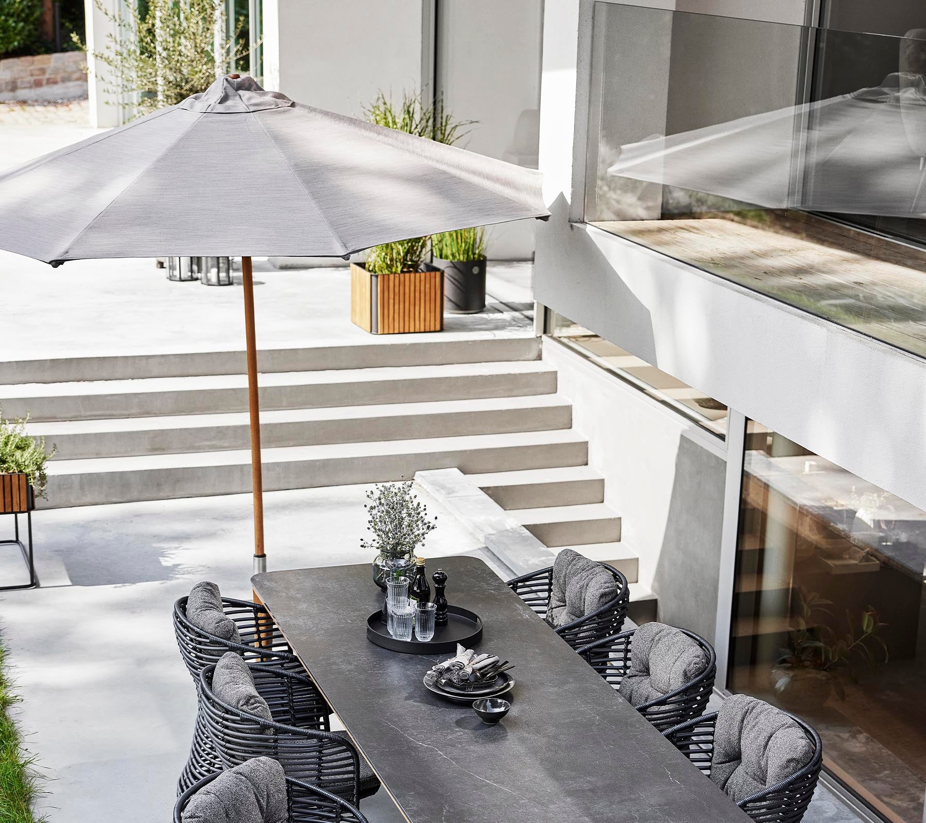 Een stijlvolle buiten eetopstelling met een lange tafel, rotan stoelen en een klassieke parasol voor schaduw.