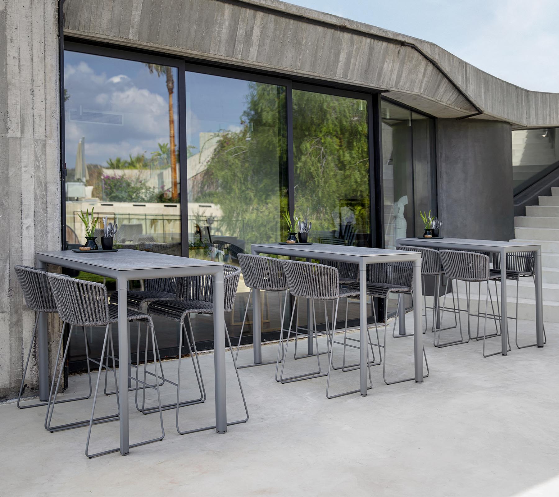 Moderne zithoek met grijze tafels en stoelen, met grote ramen die het groen buiten weerspiegelen. Minimalistische designesthetiek.