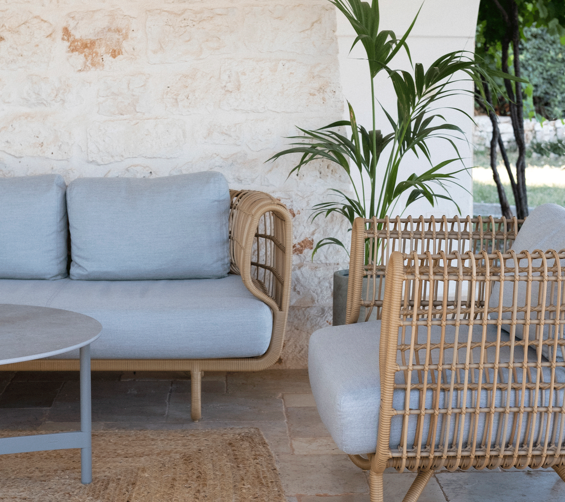 Gezellige zithoek met een lichtgrijze bank, geweven fauteuils, een ronde tafel en groen in de achtergrond. Geschikt voor ontspanning.