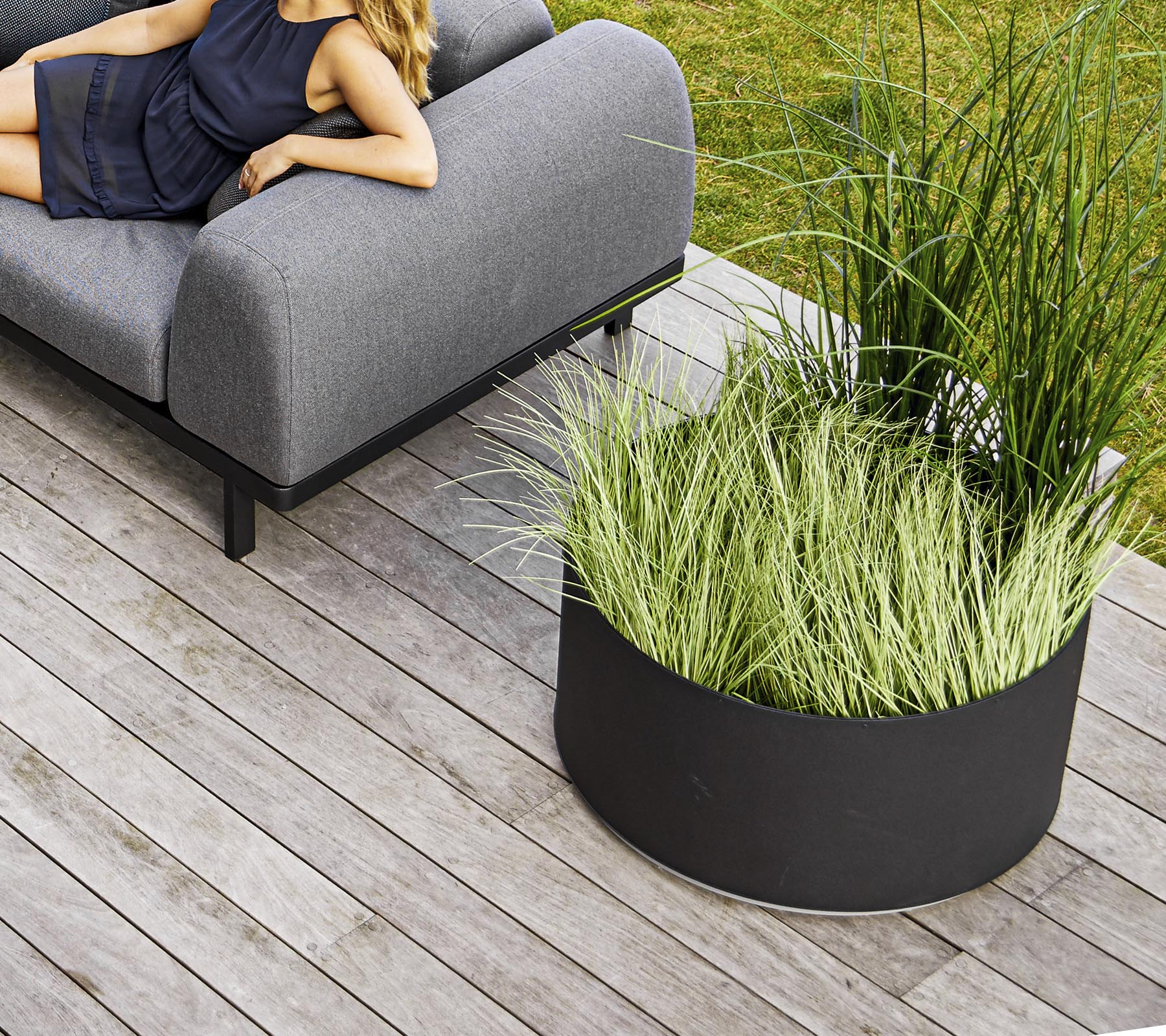 Een stijlvolle grijze bank naast een ronde zwarte plantenbak gevuld met hoog groen gras, geplaatst op houten veranda.