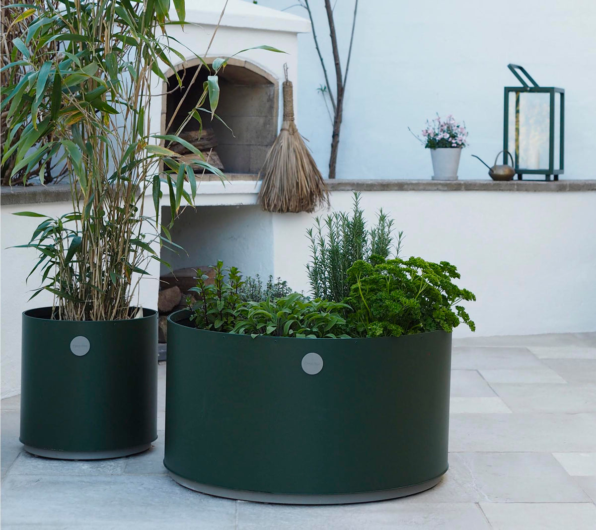 Twee ronde plantenbakken in donkergroen, de ene met hoge planten en de andere gevuld met verschillende kruiden, staan op een lichte stenen ondergrond.
