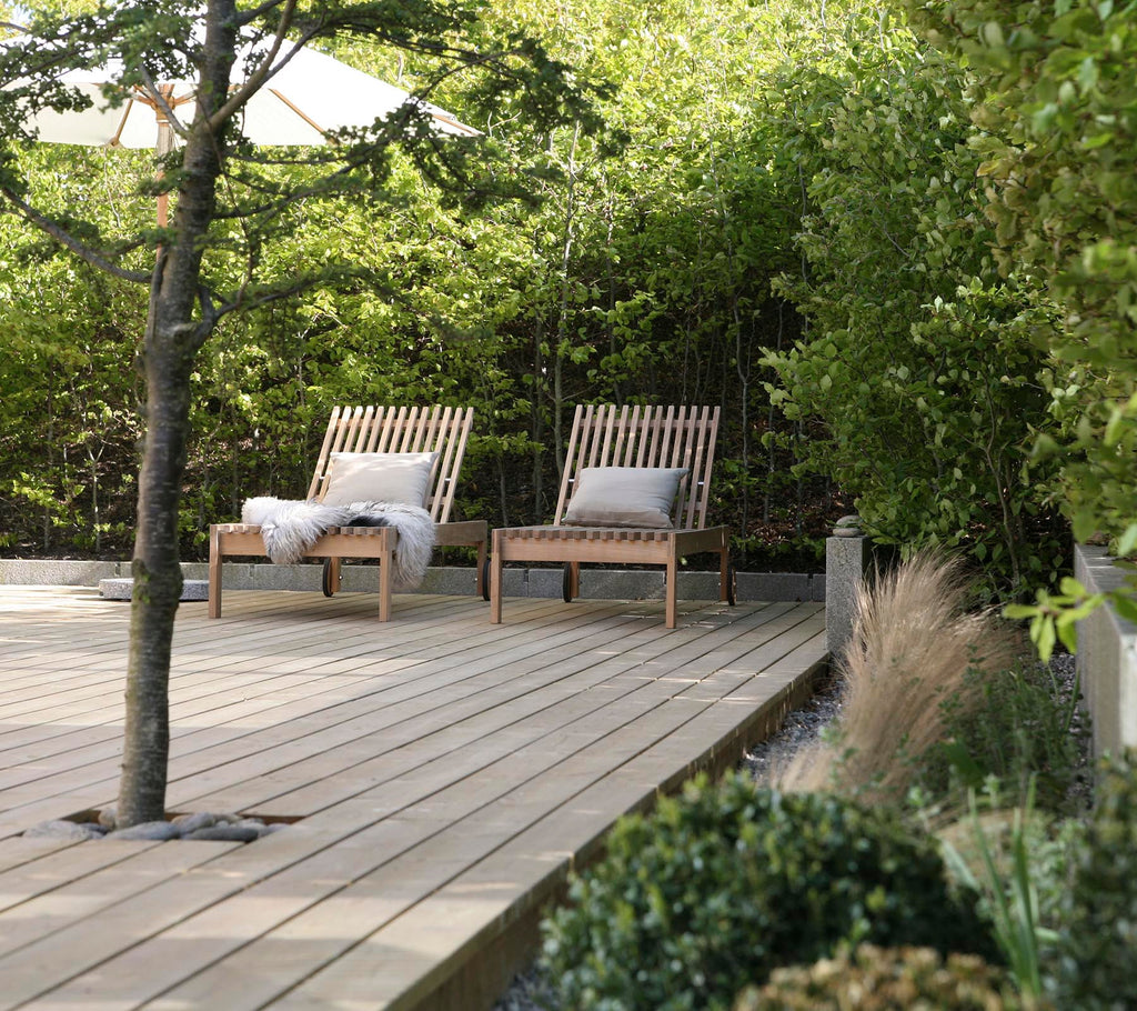 Twee houten lounge stoelen met kussens staan op een houten terras, omringd door weelderig groen en decoratieve planten.