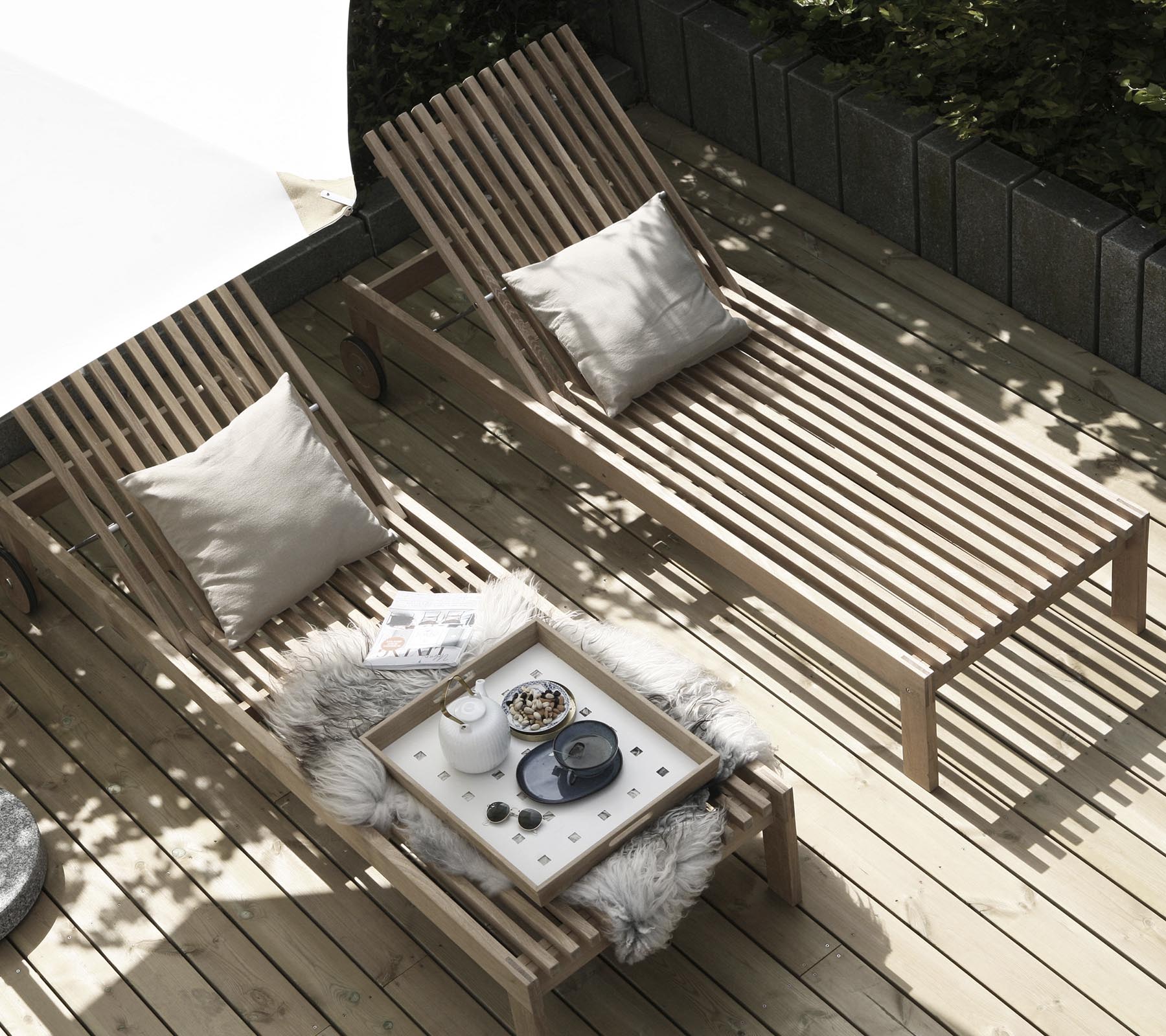 Twee houten ligstoelen met kussens zijn gerangschikt naast een klein tafeltje versierd met een gezellig dekentje en decoratieve items. Natuurlijk licht stroomt binnen.