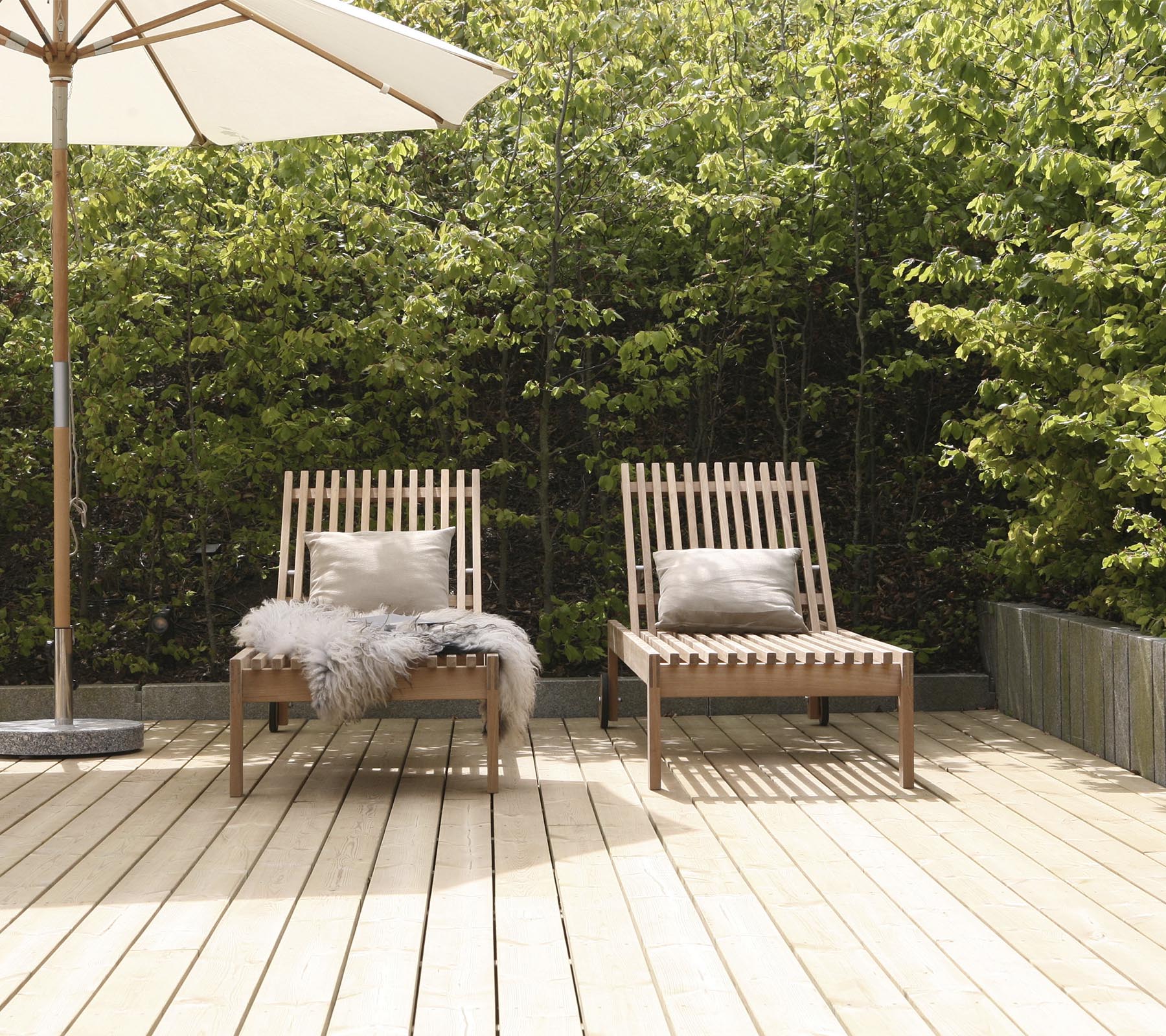 Twee stijlvolle houten lounge stoelen met kussens, geplaatst op een houten terras, omringd door groen en een grote parasol in de buurt.