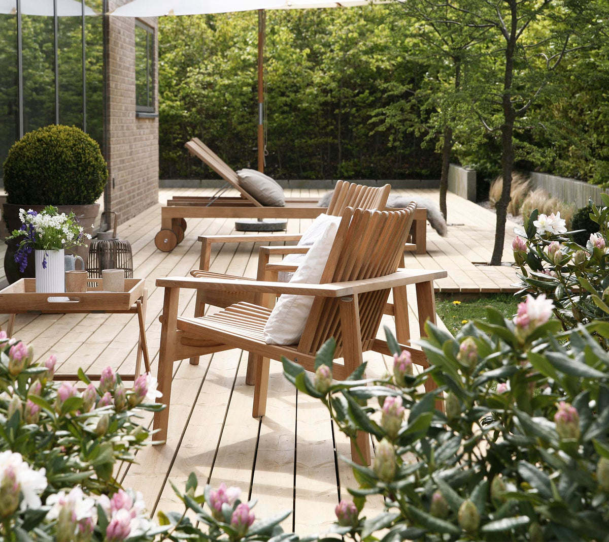 Houten stoelen en een tafel zijn omringd door groen en bloemen, wat een serene en uitnodigende buitenzitplaats creëert.
