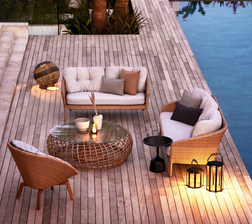 Stijlvol zitgedeelte met geweven meubels, een glazen tafel en decoratieve lampen, omgeven door hout en reflecterend water.