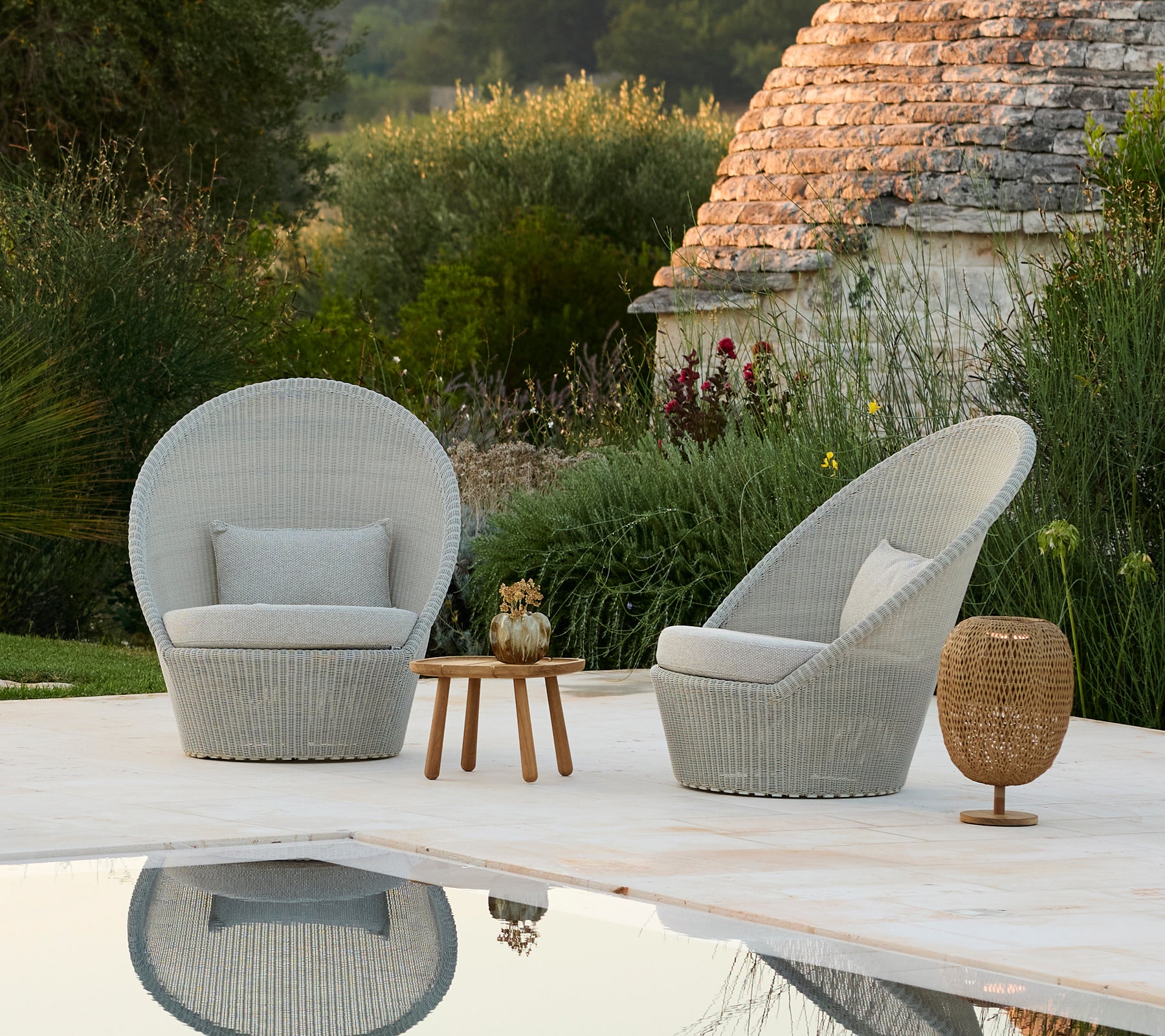 Twee grijze lounge stoelen en een houten bijzettafel naast een zwembad in een moderne buitenomgeving.
