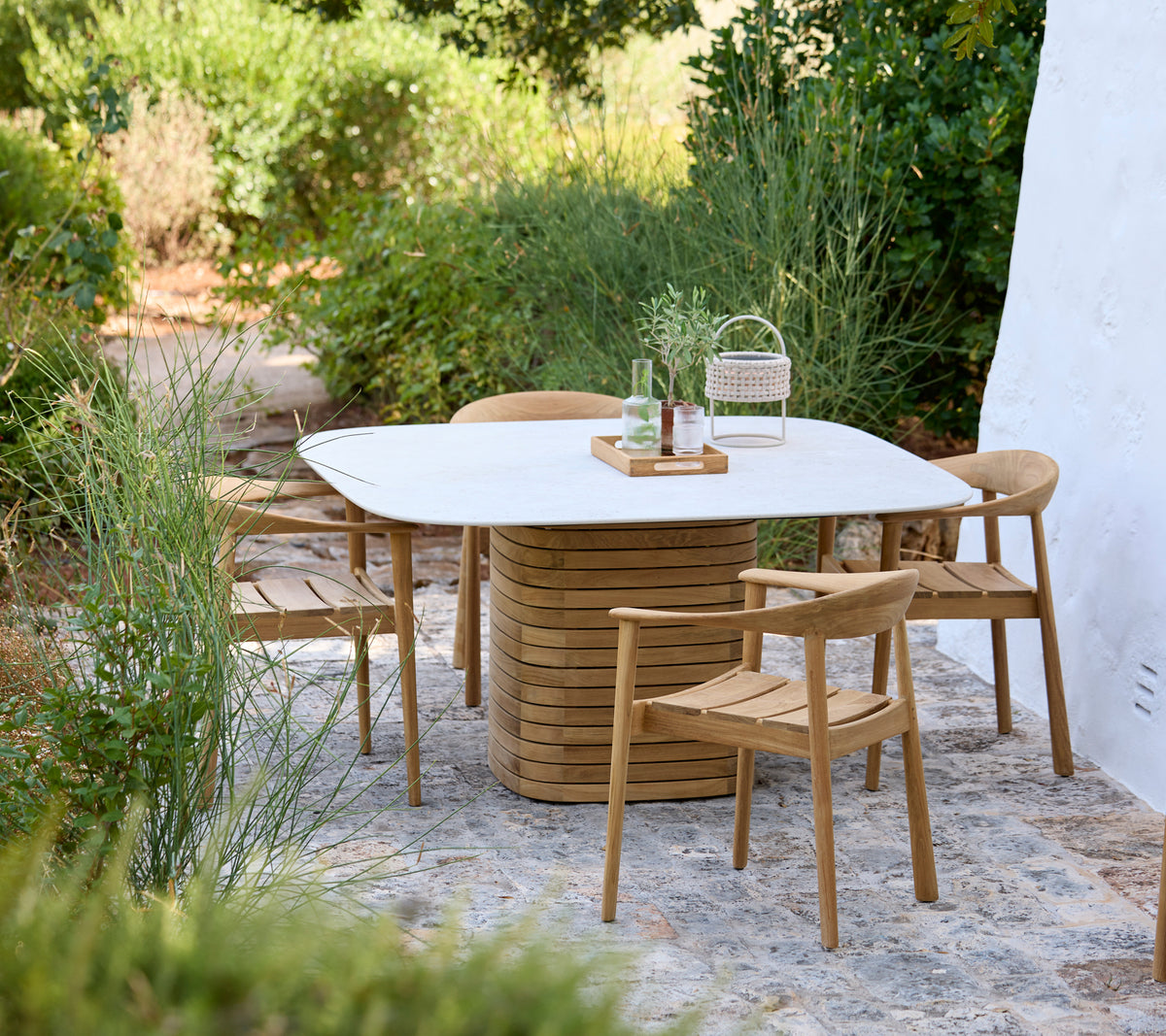 Moderne buitenruimte met een witte tafel en houten stoelen omringd door groen.
