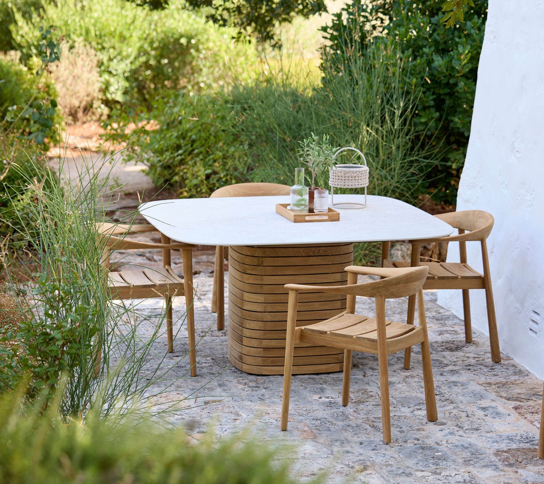 Moderne buitenruimte met een witte tafel en houten stoelen omringd door groen.