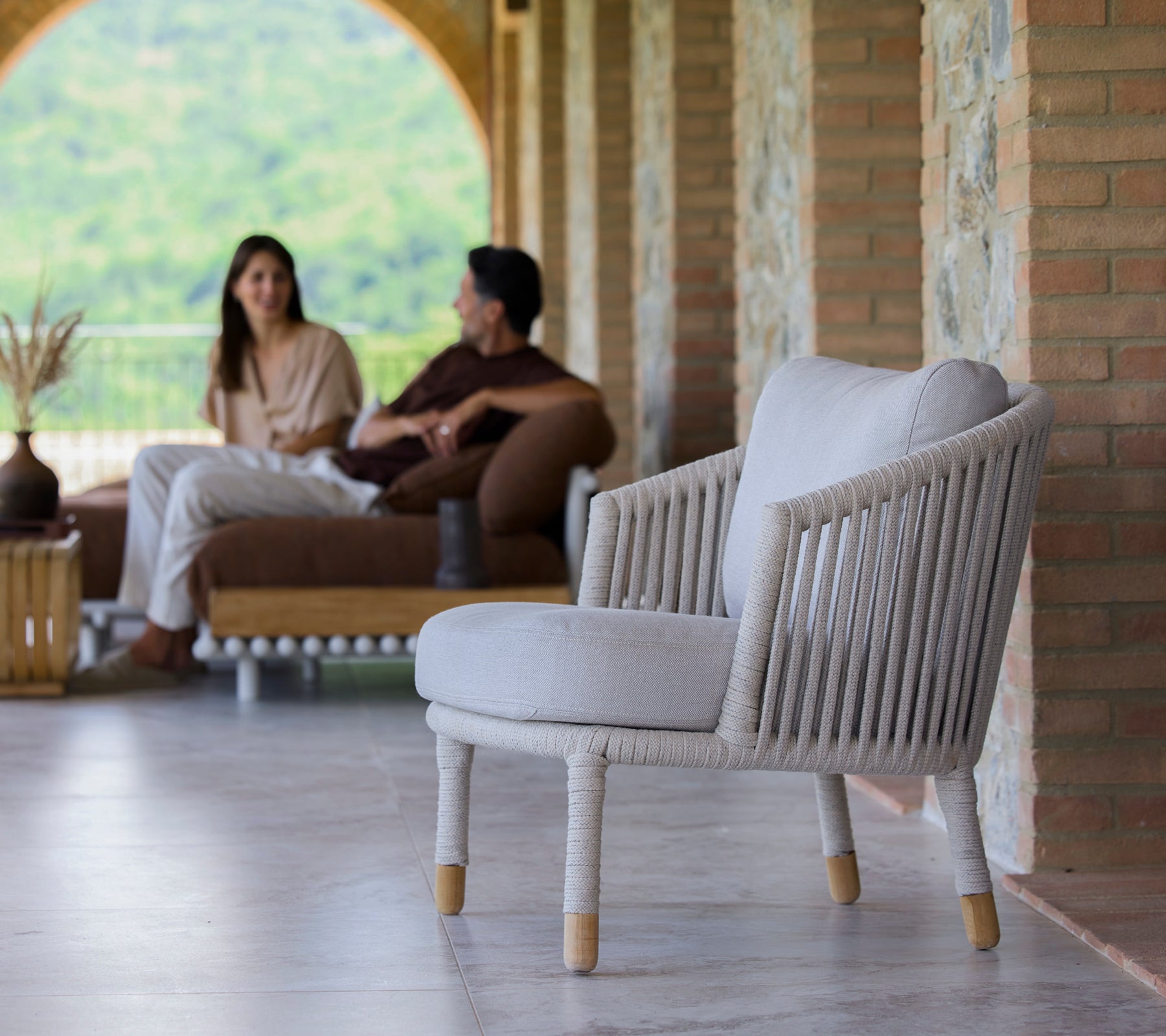 Een zandkleurige lounge fauteuil met een minimalistisch design, weergegeven in een serene omgeving met mensen die in gesprek zijn.