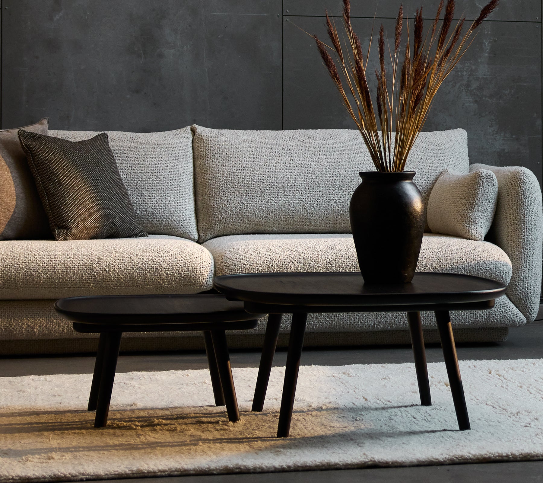 Twee elegante zwarte salontafels in verschillende maten, aangevuld met een decoratieve vaas met gedroogde planten, geplaatst tegenover een gezellige bank.
