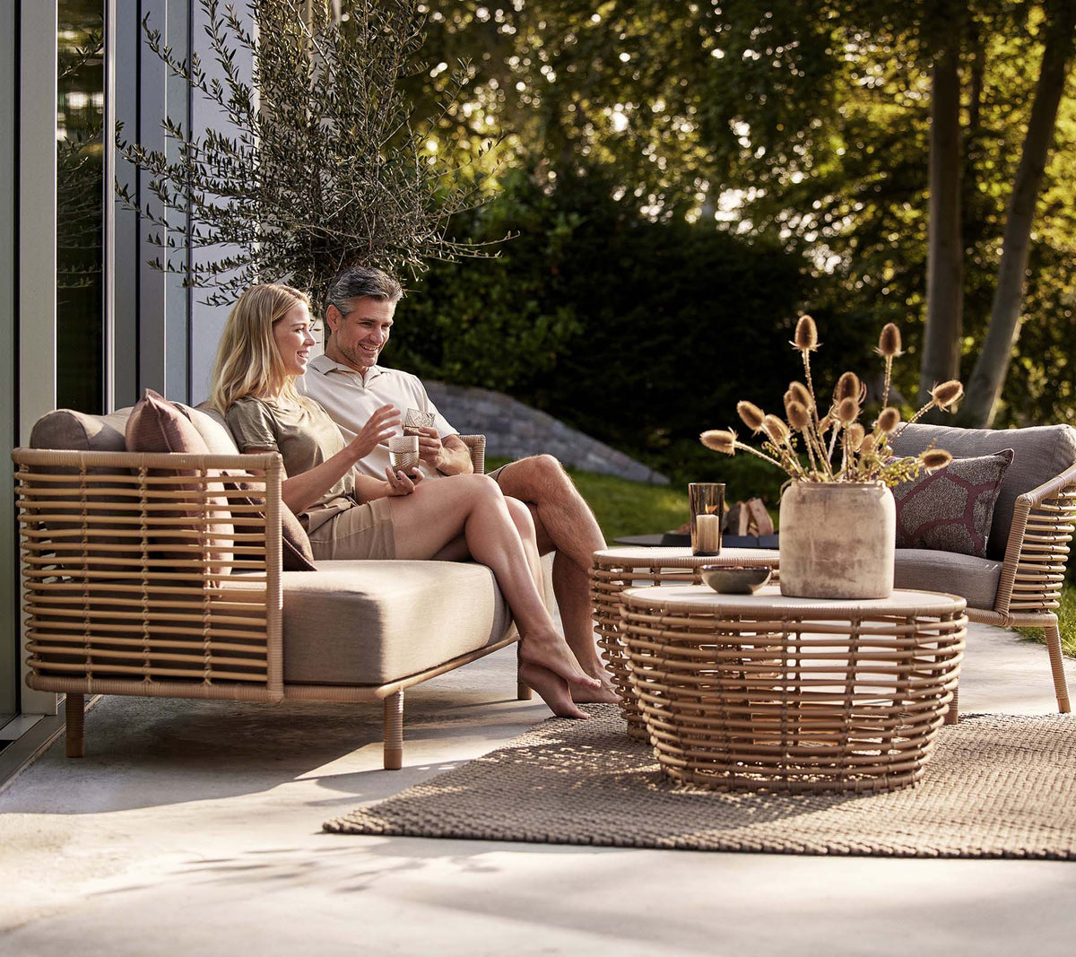 Een stel geniet van drankjes op een Sense-sofa en lounge stoel, vergezeld van een stijlvolle mand koffie tafel en een decoratieve vaas.
