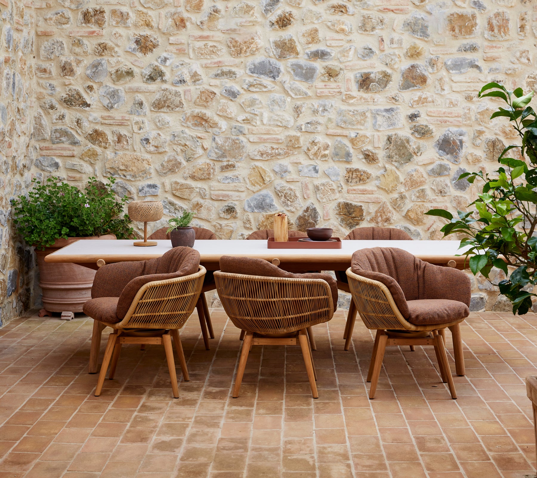 Een houten eettafel omringd door stijlvolle stoelen met teak poten, geplaatst tegen een gestructureerde stenen muur en versierd met groene planten.