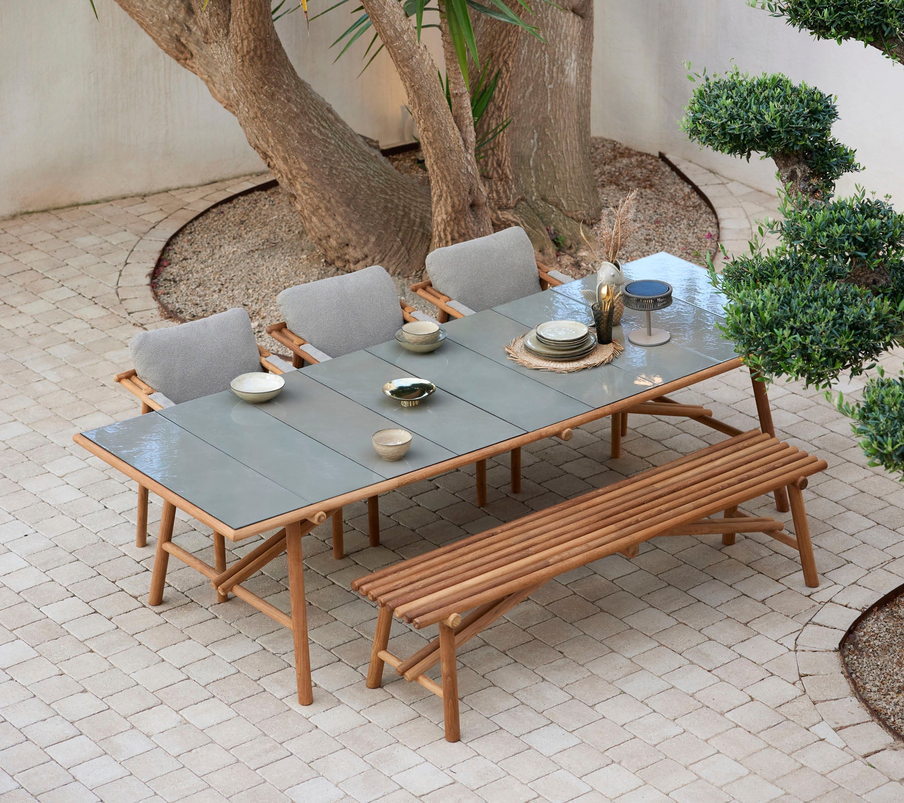 Een houten tafel gedekt met borden en glazen, omringd door comfortabele stoelen en een bank, alles geplaatst tussen het groen.