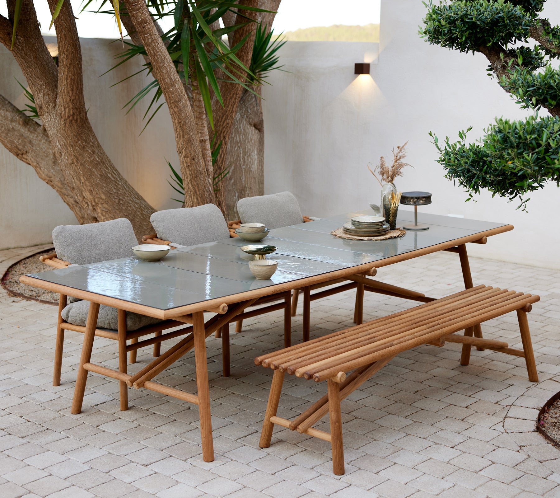 Teakhouten eettafel met glazen blad, vergezeld van een bank en vier beklede stoelen, gedekt met servies en sfeervolle verlichting.