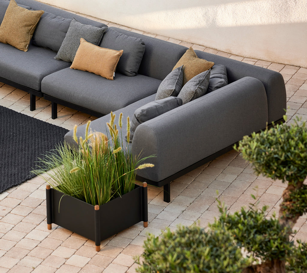 Lava grijze sofa met decoratieve kussens naast een moderne takken plantenbak gevuld met weelderig groen, geplaatst op een gestructureerd oppervlak.