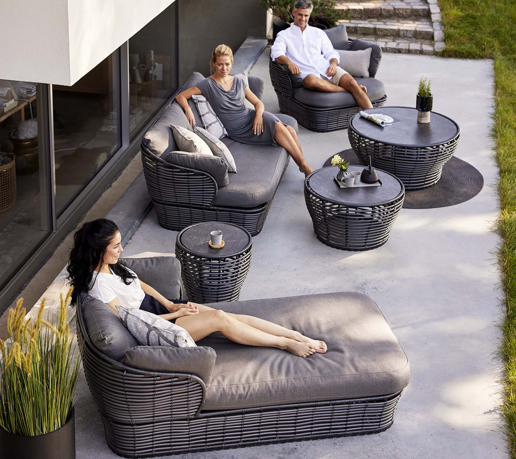 Moderne lounge-ruimte met een mand-dagbed, een 3-zitsbank en loungesstoelen, aangevuld met ronde tafels en decoratieve planten.