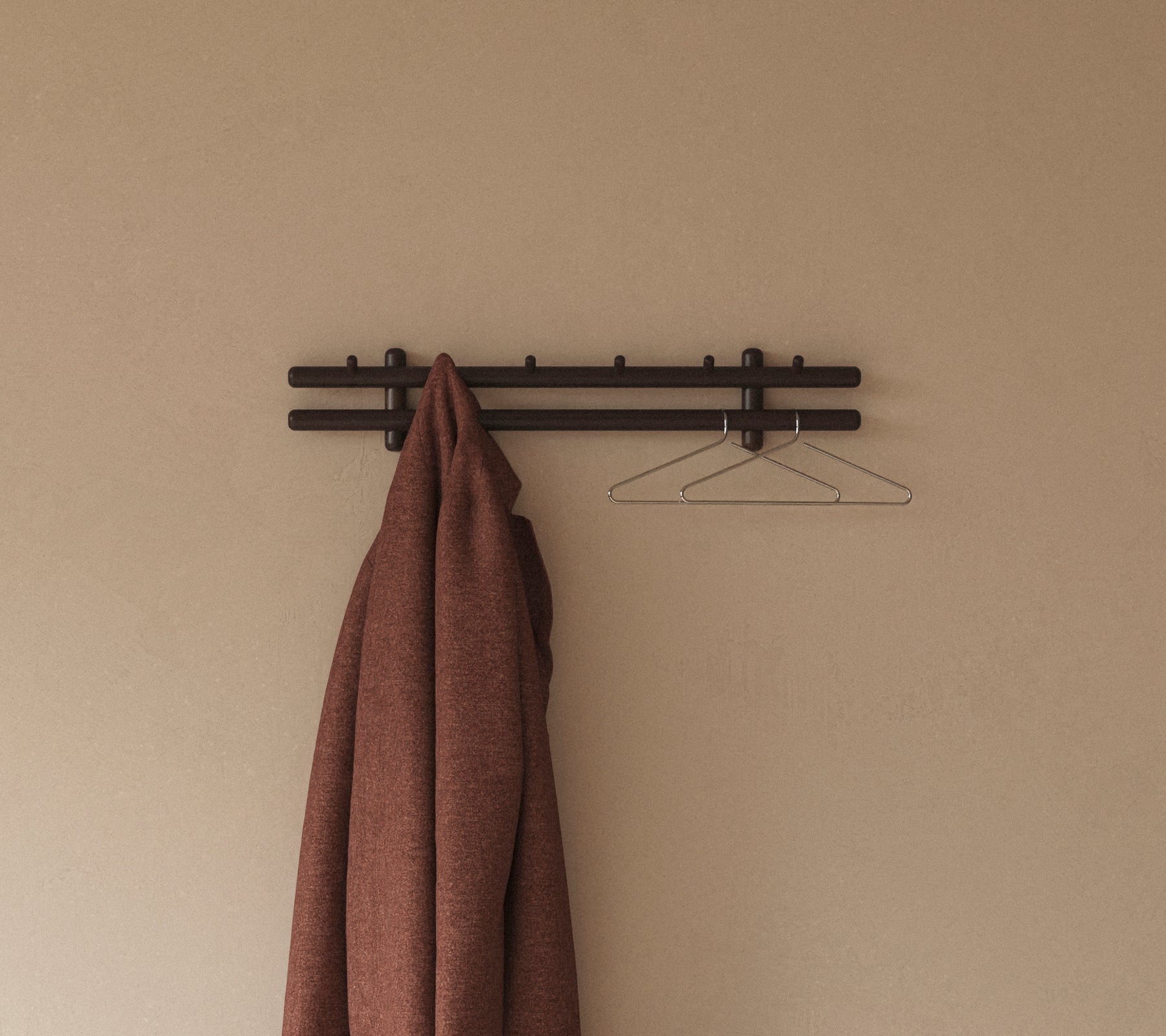 Een bruine jas hangt aan een minimalistische wandrek met haken, aangevuld door een eenvoudige draadhanger tegen een neutrale achtergrond.