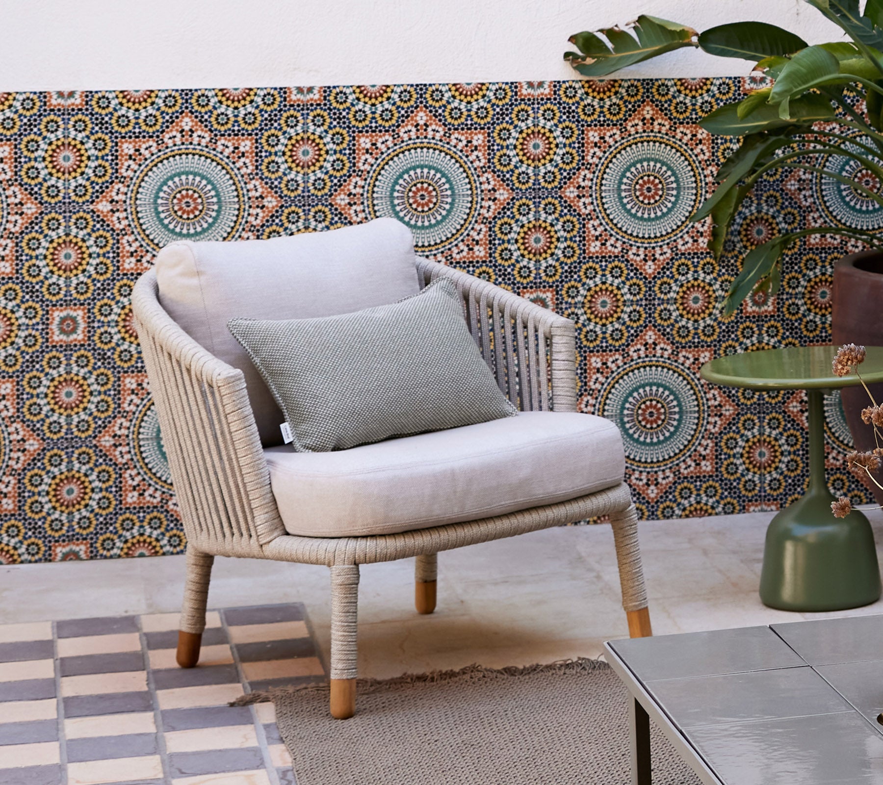 Een stijlvolle loungechaise met een zachte kussen en een decoratiekussen, geplaatst tegen een gemusterde achtergrond en omringd door een potplant.