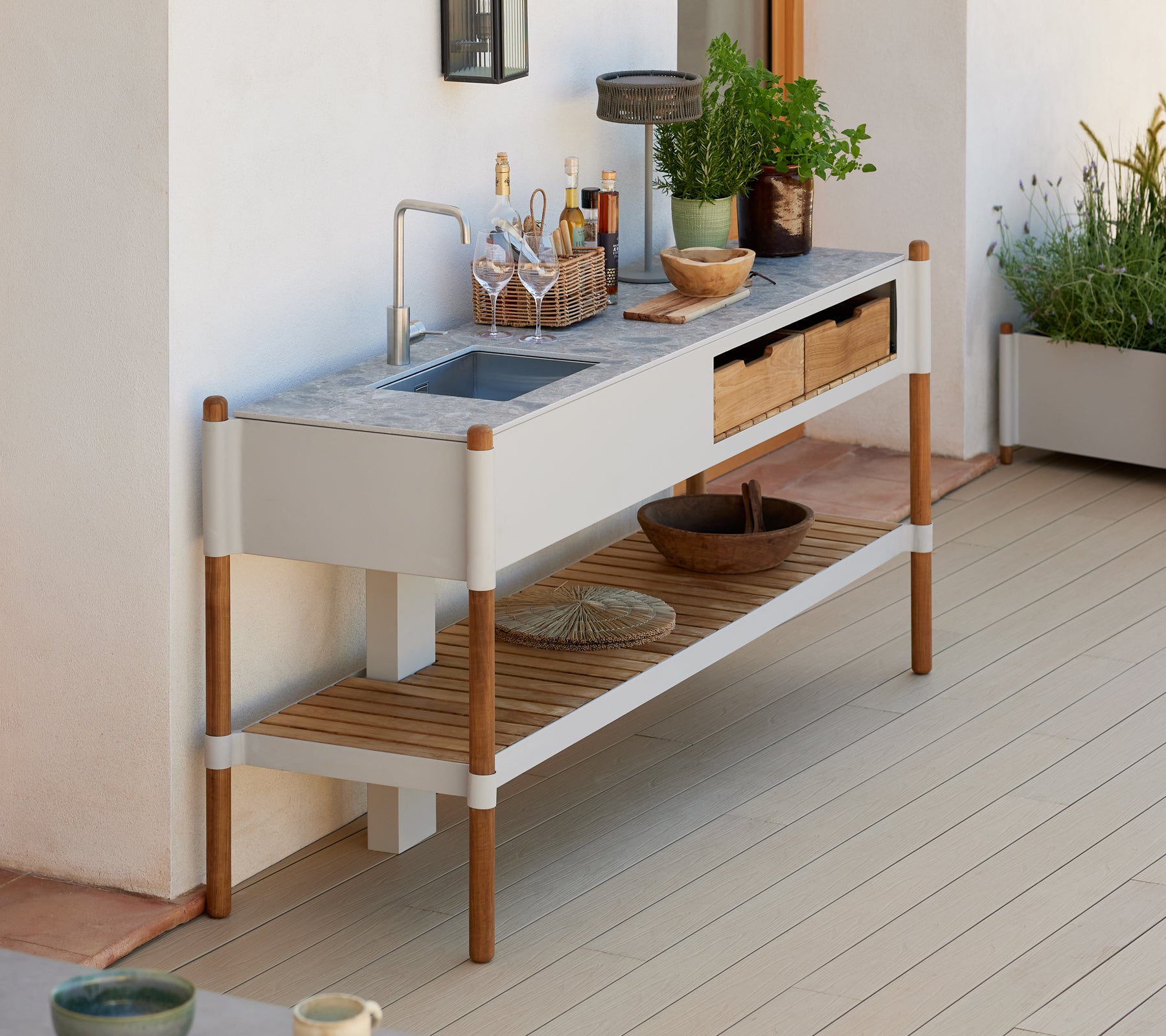 Een moderne, minimalistische buitensinkstation met een granieten aanrechtblad, houten opbergladen en decoratieve plantenarrangementen.
